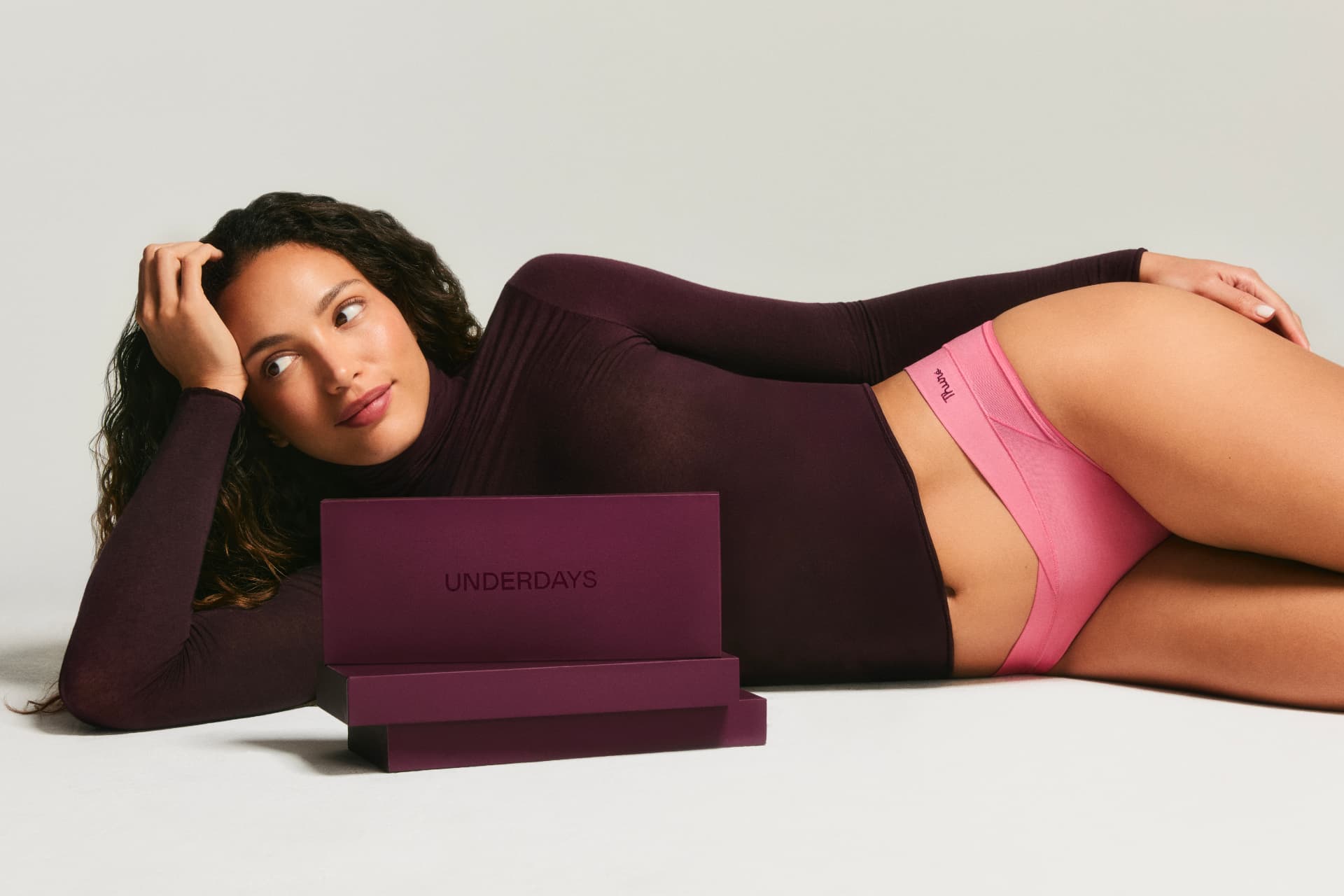 Model reclining beside an Underdays gift box wearing pink briefs
